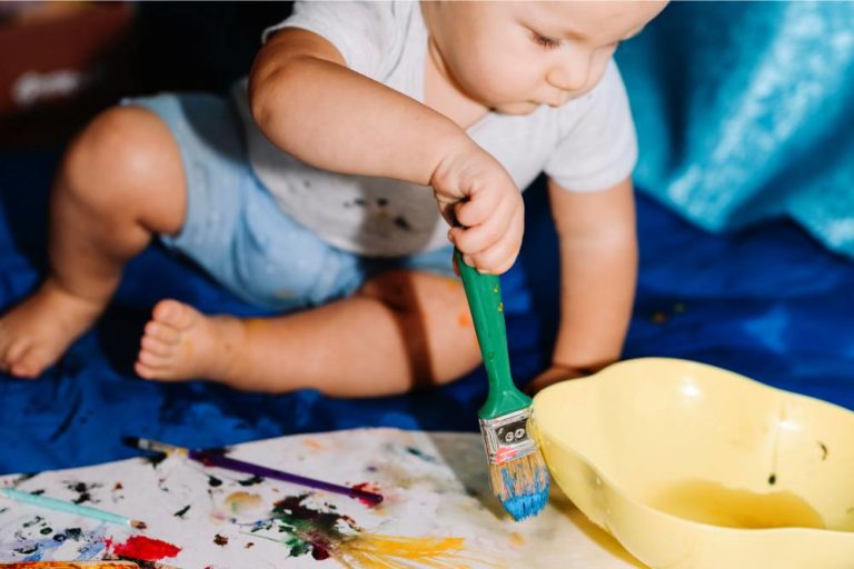 Criança sentada sobre panos, pintando um cartaz branco sobre o chão usando tinta e pincel.